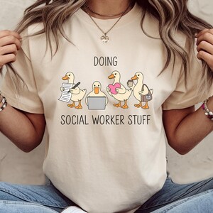 May include: Beige t-shirt with the text "DOING SOCIAL WORKER STUFF" above a graphic of four ducks. The ducks are depicted with various items, including a pen and paper, a laptop, a heart, a coffee cup, and a briefcase.