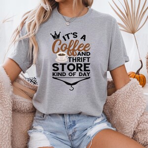 May include: A gray t-shirt with a graphic design that says "It's a Coffee and Thrift Store Kind of Day". The graphic includes a crown, a coffee cup, and a hanger.