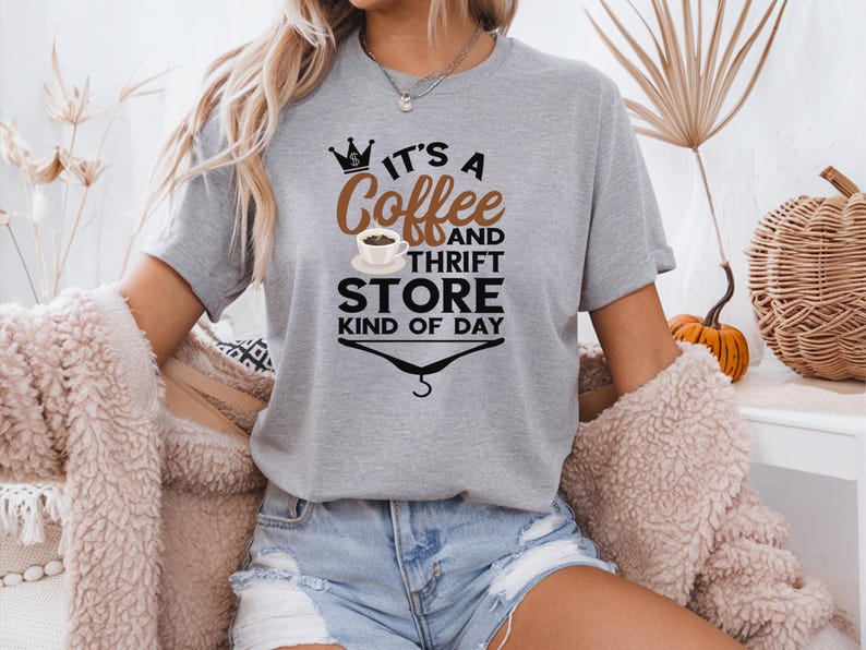 May include: A gray t-shirt with a graphic design that says "It's a Coffee and Thrift Store Kind of Day". The graphic includes a crown, a coffee cup, and a hanger.