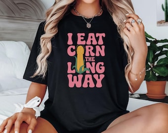 May include: Black t-shirt with the text "I EAT CORN THE LONG WAY" in pink, retro-style lettering. A yellow corn graphic is centered between the words. The shirt is a casual, everyday wear item.