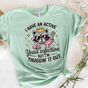 May include: A light green t-shirt with a cartoon raccoon wearing a crown and sunglasses. The text on the shirt reads "I have an active yeast infection but I'm thuggin' it out."