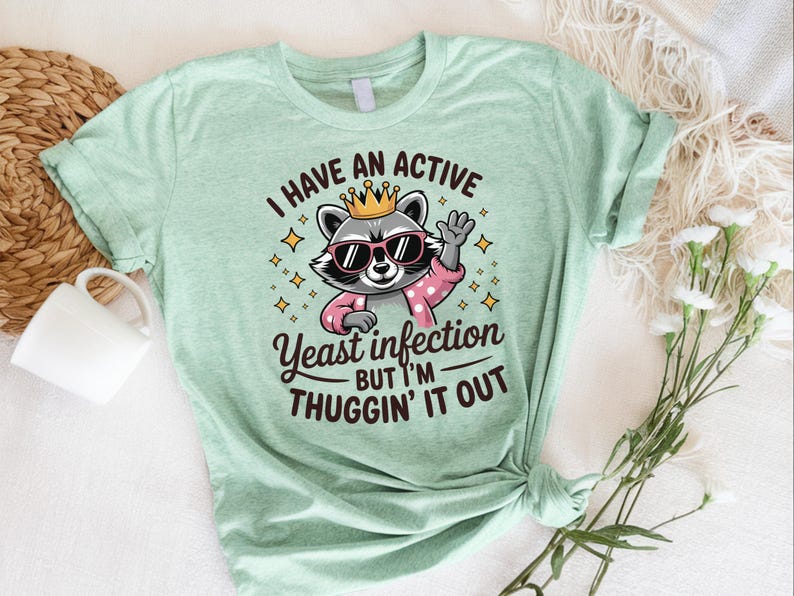 May include: A light green t-shirt with a cartoon raccoon wearing a crown and sunglasses. The text on the shirt reads "I have an active yeast infection but I'm thuggin' it out."