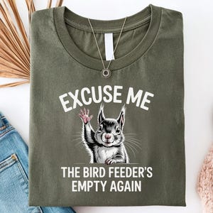 May include: Olive green t-shirt with the text "EXCUSE ME" above a graphic of a squirrel waving. Below the squirrel is the text "THE BIRD FEEDER'S EMPTY AGAIN". The shirt is folded with a necklace and jeans visible.