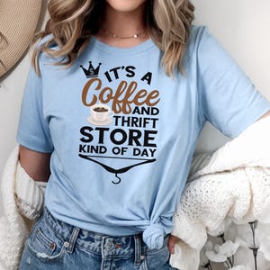 May include: A light blue t-shirt with a graphic design that says "It's a Coffee and Thrift Store Kind of Day". The graphic includes a crown, a coffee cup, and a hanger.