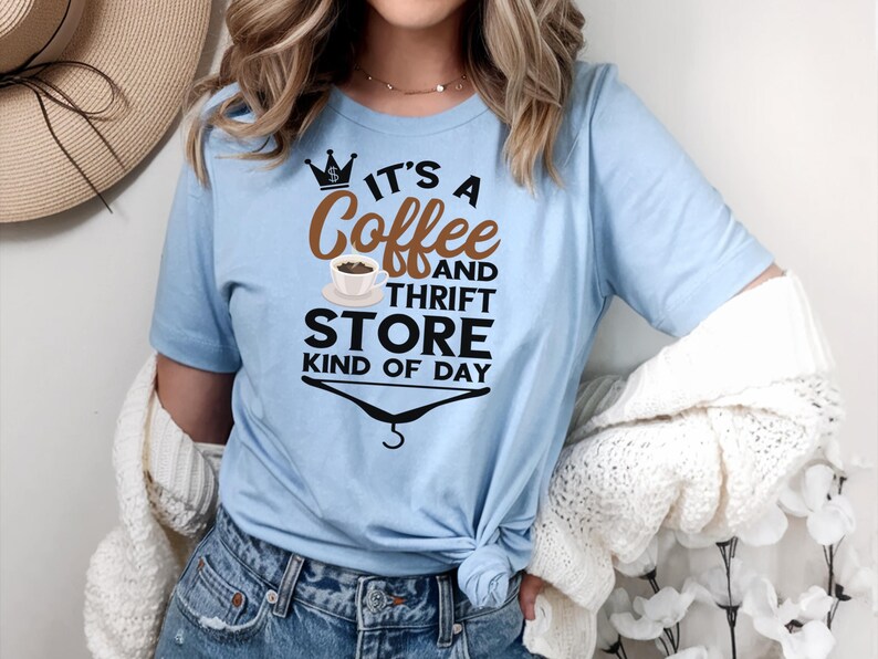 May include: A light blue t-shirt with a graphic design that says "It's a Coffee and Thrift Store Kind of Day". The graphic includes a crown, a coffee cup, and a hanger.