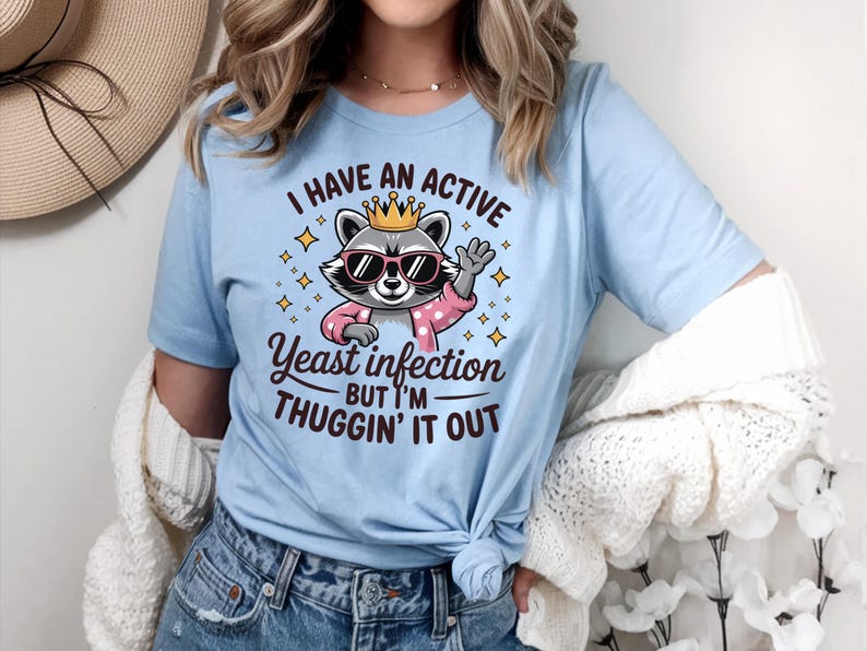 May include: Light blue t-shirt with a cartoon raccoon wearing a crown and sunglasses. The text on the shirt reads "I have an active yeast infection but I'm thuggin' it out."