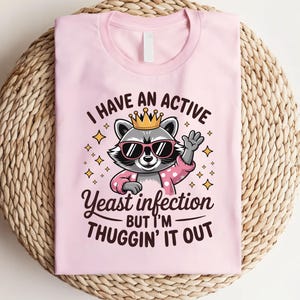 May include: Pink t-shirt with a cartoon raccoon wearing a crown and sunglasses. The text on the shirt reads "I have an active yeast infection but I'm thuggin' it out."