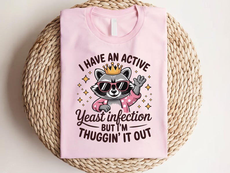 May include: Pink t-shirt with a cartoon raccoon wearing a crown and sunglasses. The text on the shirt reads "I have an active yeast infection but I'm thuggin' it out."