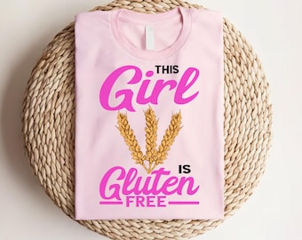 May include: A light pink t-shirt with the text "THIS Girl IS Gluten FREE" in bright pink. The design includes three golden wheat stalks. The shirt is displayed on a woven, circular surface.