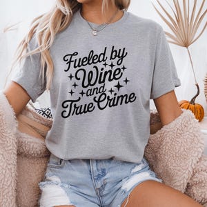 May include: A gray t-shirt with the text "Fueled by Wine and True Crime" in black lettering with stars around the words.