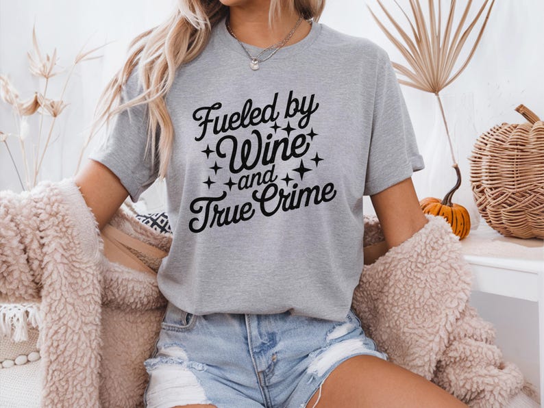 May include: A gray t-shirt with the text "Fueled by Wine and True Crime" in black lettering with stars around the words.