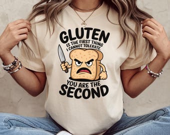 May include: A beige t-shirt with a graphic of a slice of bread with a knife and the text "Gluten is the first thing I cannot tolerate. You are the second."