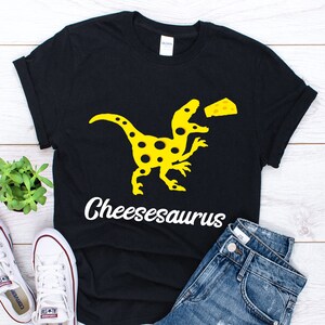Könnte beinhalten: Schwarzes T-Shirt mit einem gelben Cheeseburger-Dinosaurier-Motiv und dem Text "Cheesesaurus".