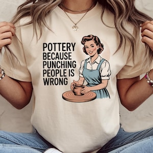 May include: Beige t-shirt with the text "Pottery Because Punching People Is Wrong" and a vintage illustration of a woman working on a pottery wheel. The woman wears a blue apron and a white shirt.