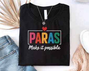May include: Black t-shirt with the word "PARAS" in colorful letters inside a white box, topped with a red heart. Below, the phrase "Make it possible" is written in white. A silver necklace is visible.