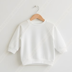 May include: A plain white long-sleeved sweatshirt for children, hanging on a wooden hanger. The sweatshirt has a crew neck and raglan sleeves. The text "INCLUDED IN YOUR PURCHASE" is visible on the sides.