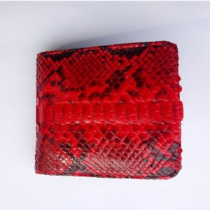 May include: A red bi-fold wallet with a snakeskin pattern. The wallet is made of a textured material with a repeating scale design. The color is a vibrant red with black accents, creating a bold and eye-catching accessory.