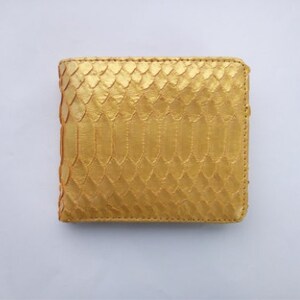 May include: A gold-colored, square wallet with a textured, scaly pattern. The wallet is made of a material that resembles snakeskin. The edges are stitched with a matching gold thread. The wallet is closed and appears new.