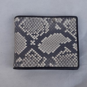 May include: A bi-fold wallet with a snakeskin pattern in shades of gray and black. The wallet has a black border and a textured surface. The design features a repeating pattern of scales.