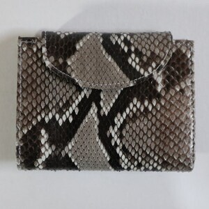May include: A small, rectangular wallet with a flap closure. The wallet is made of a snakeskin-patterned material in shades of gray, brown, and white. The flap is rounded and secured with a snap. The stitching is white.