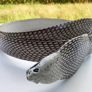 May include: A coiled belt made from snake skin, featuring a detailed pattern of scales in shades of grey and white. The belt is designed with a snake head buckle, adding a unique and exotic touch to the accessory.