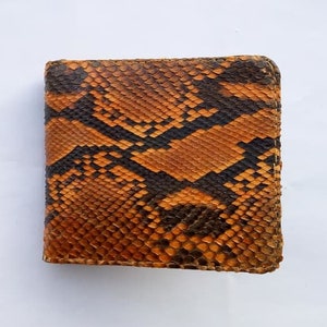 May include: A square, bi-fold wallet made of genuine snakeskin. The wallet features a pattern of brown and black scales. The edges are stitched with a matching thread. The wallet is shown on a white background.