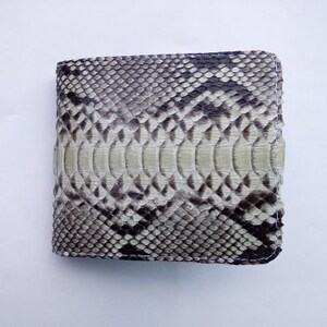 May include: A bi-fold wallet with a snakeskin pattern in shades of gray, black, and cream. The wallet is square-shaped and appears to be made of genuine leather. The stitching is white.