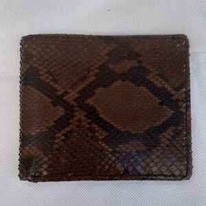 Peut inclure: Un portefeuille marron à deux volets avec un motif peau de serpent. Le portefeuille est rectangulaire et semble être en cuir. Le motif présente des nuances de marron et de noir, créant un aspect texturé.