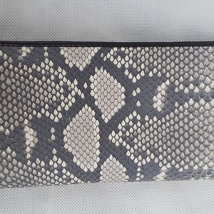 May include: A rectangular wallet with a snakeskin pattern in shades of gray and white. The wallet has a black border and a textured surface, showcasing a natural, reptile-inspired design. The overall aesthetic is sleek and sophisticated.