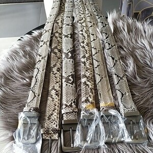 May include: A collection of six belts with a snakeskin pattern in shades of gray, black, and white. Each belt has a silver-colored buckle and is partially wrapped in clear plastic. The belts are arranged on a fluffy, gray surface.