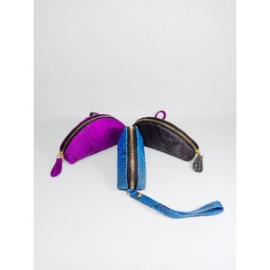 May include: Three small, semi-circular zippered pouches in vibrant colors. One is a deep purple, another is a textured black, and the third is a bright blue with a wrist strap. All have gold-toned zippers and pulls.