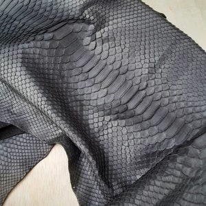 May include: Close-up of a piece of dark grey snakeskin leather. The leather has a textured, scaly pattern with variations in tone, creating a realistic appearance. The material is folded, revealing the underside and adding depth.