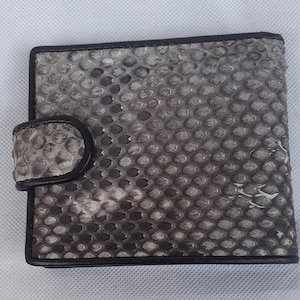 May include: A square wallet with a snakeskin pattern in shades of gray and black. The wallet has a black border and a flap with a snap closure. The texture of the snakeskin is visible.