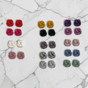 May include: A collection of twenty-two stud earrings arranged in pairs, each featuring a knotted, circular design. The earrings come in a variety of solid matte colours including red, pink, white, mustard yellow, black, grey, purple, mauve, blue, and sage green.