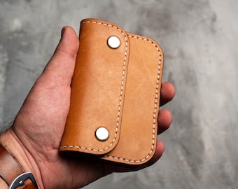 May include: A brown leather key holder with two snap closures and white stitching. The key holder is folded in half and held in a hand.
