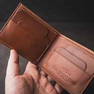 May include: A brown leather wallet with multiple card slots and a stitched edge. The wallet is open and the inside is visible, showing a brand logo that reads "Rus Leather Goods".