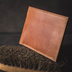 May include: A brown leather wallet with stitching around the edges, resting on a brown brush with dark bristles.