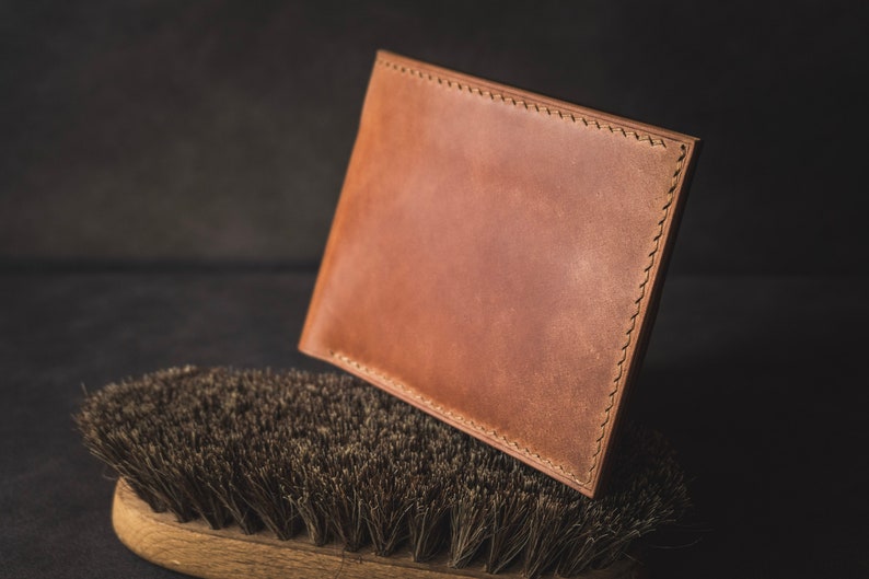May include: A brown leather wallet with stitching around the edges, resting on a brown brush with dark bristles.