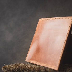 May include: A light brown leather wallet with stitching around the edges, resting on a brown bristled brush.