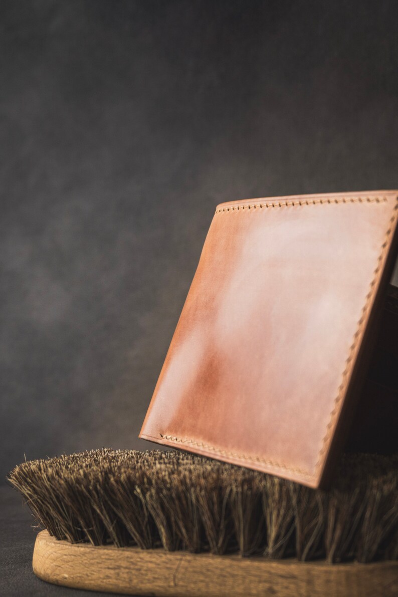 May include: A light brown leather wallet with stitching around the edges, resting on a brown bristled brush.