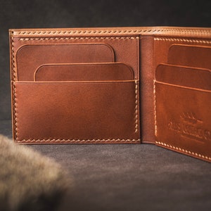 May include: A brown leather wallet with multiple card slots and a large bill compartment. The wallet is open and the inside is visible. The wallet has a stitched edge and a logo that says "Old World Goods".
