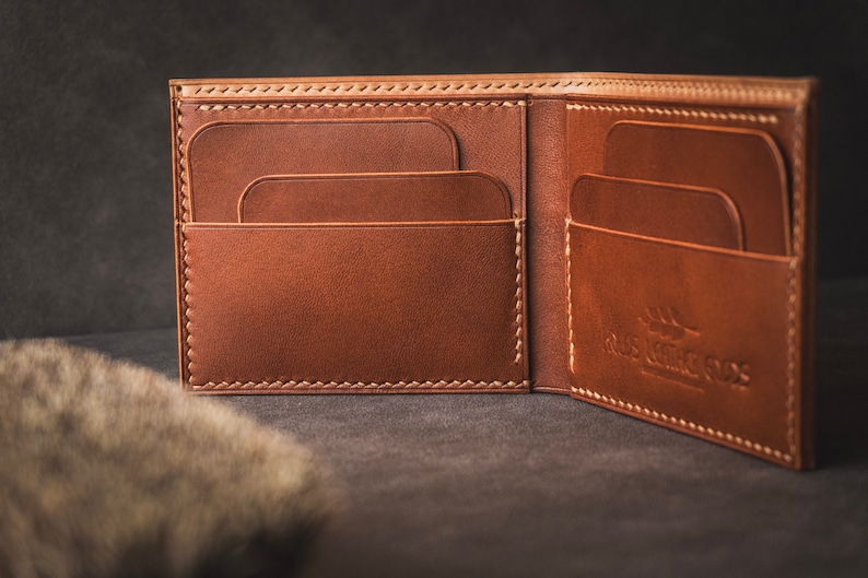 May include: A brown leather wallet with multiple card slots and a large bill compartment. The wallet is open and the inside is visible. The wallet has a stitched edge and a logo that says "Old World Goods".