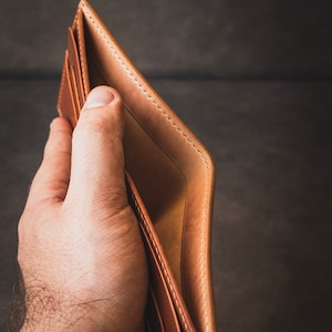 May include: A brown leather wallet with stitching detail is open and held in a hand. The wallet is empty.