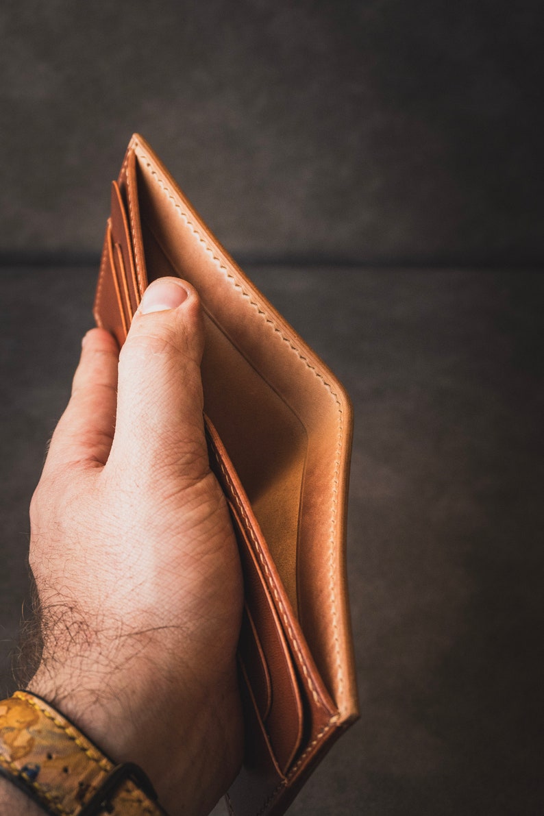 May include: A brown leather wallet with stitching detail is open and held in a hand. The wallet is empty.