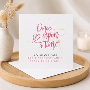 K&ouml;nnte beinhalten: Eine wei&szlig;e Karte mit rosa Schriftzug "Once Upon a Time". Darunter steht "A wish was made and a forever family began their story." Die Karte liegt auf einem Holztablett, mit einer Vase und einer Kerze im Hintergrund.