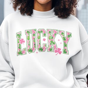 May include: White sweatshirt with the word "LUCKY" in green and pink floral lettering. The letters are outlined in green and feature shamrock and star details. The sweatshirt has a high neckline.