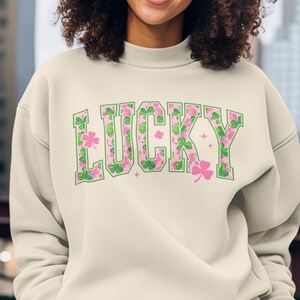 May include: Cream-colored sweatshirt with the word "LUCKY" in green outlined letters filled with pink and green shamrock patterns. The sweatshirt has a crew neck and long sleeves, perfect for St. Patrick's Day.
