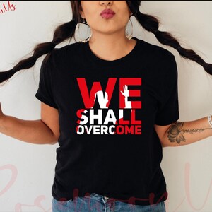 May include: Black t-shirt with the words "WE SHALL OVERCOME" in white letters with a red outline. The words are in a bold, blocky font. A white silhouette of a raised fist is in the middle of the "WE" part of the text.
