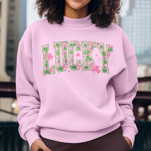 May include: A light pink sweatshirt with the word "LUCKY" in green outlined letters. The letters are filled with a pattern of pink and green shamrocks. The sweatshirt has a crew neck and long sleeves.