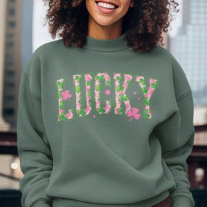 May include: A green sweatshirt with the word "LUCKY" in large, floral letters. The letters are filled with a pattern of pink and green shamrocks. A pink shamrock is on the right side of the word. The sweatshirt has a high neckline.